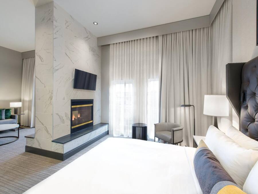 A modern hotel room with a large bed, a fireplace, a television, and a view of the city through large windows.