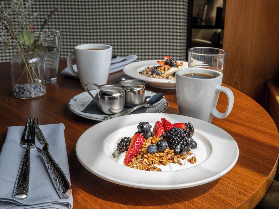 A breakfast setting with a plate of yogurt topped with berries and granola, a cup of coffee, and a glass of water on a wooden table.