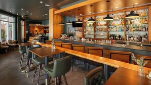A bar area with wooden tables and chairs, a bar counter with shelves of bottles, and a large window providing natural light.