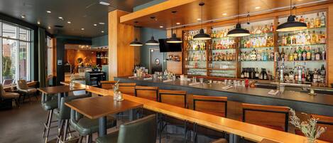 A bar area with wooden tables and chairs, a bar counter with shelves of bottles, and a large window providing natural light.