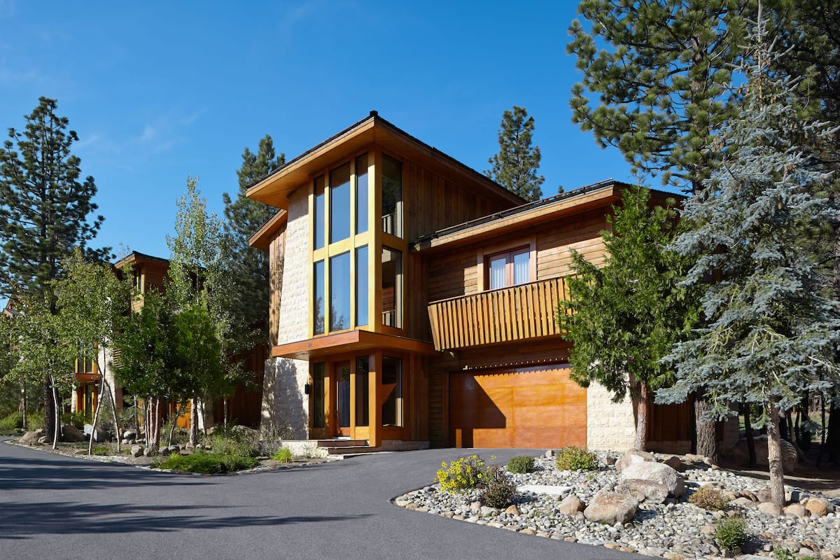 A modern two-story house with a large balcony, surrounded by trees and a stone pathway.