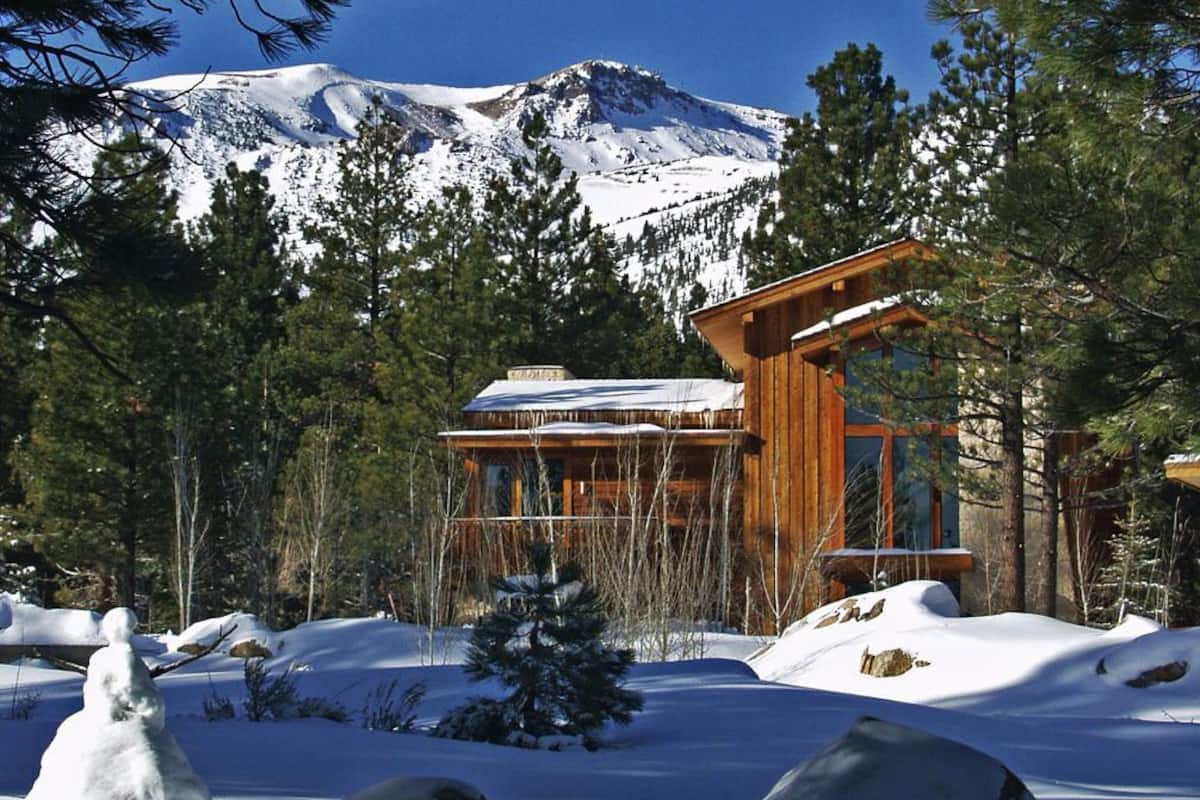A wooden cabin with a snow-covered roof, surrounded by snow and trees, with a mountain in the background.