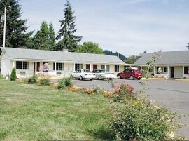 Um motel com estrutura em um único pavimento, área de estacionamento coberta e um pequeno jardim com flores.