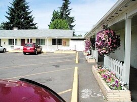 Um motel com estacionamento, um carro vermelho estacionado na frente e um canteiro de flores com flores rosa e brancas no lado direito.