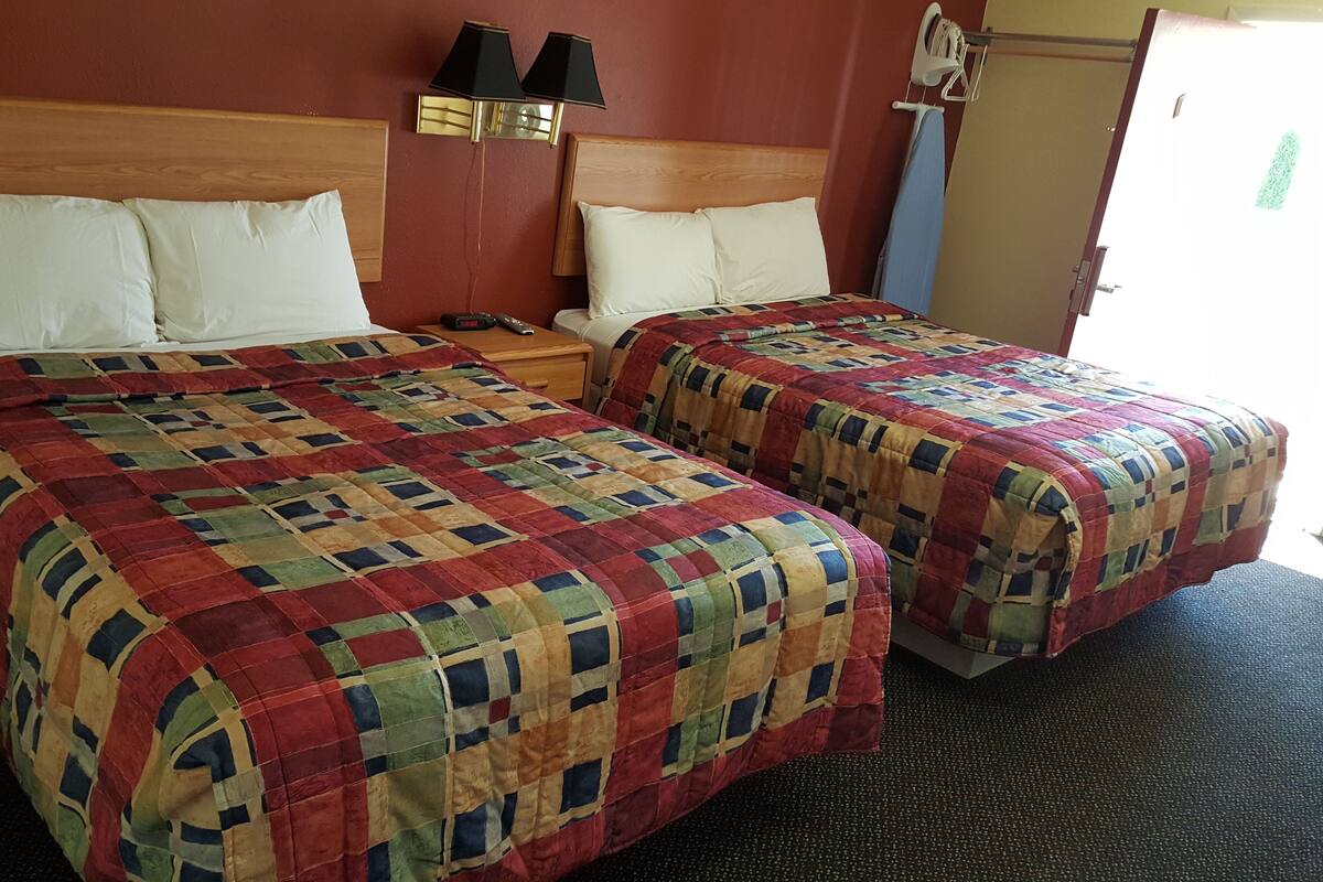 A hotel room with two beds, each with a quilted bedspread, a bedside table with a lamp, and a wall-mounted light fixture.
