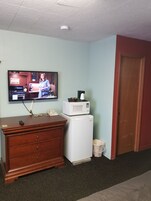 Quarto com cômoda de madeira, geladeira branca, micro-ondas, televisão e porta.