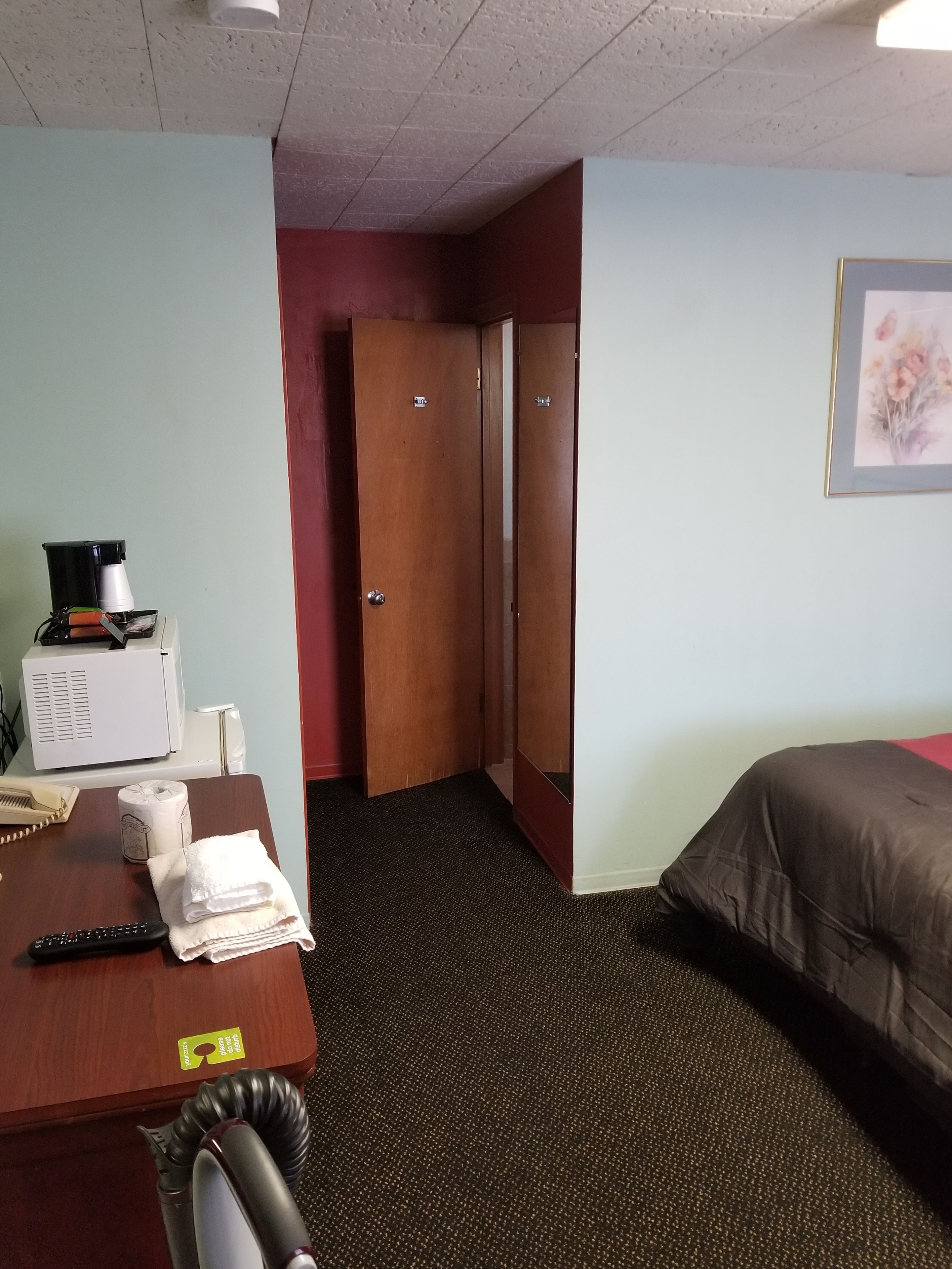 A hotel room with a desk, a bed, a wall-mounted picture, and a door leading to another room.