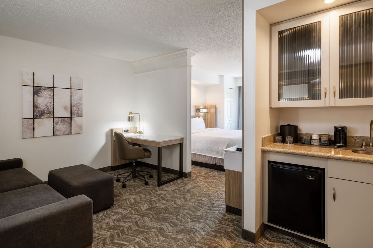 A hotel room with a bed, a desk, a chair, a kitchenette, and a small refrigerator.