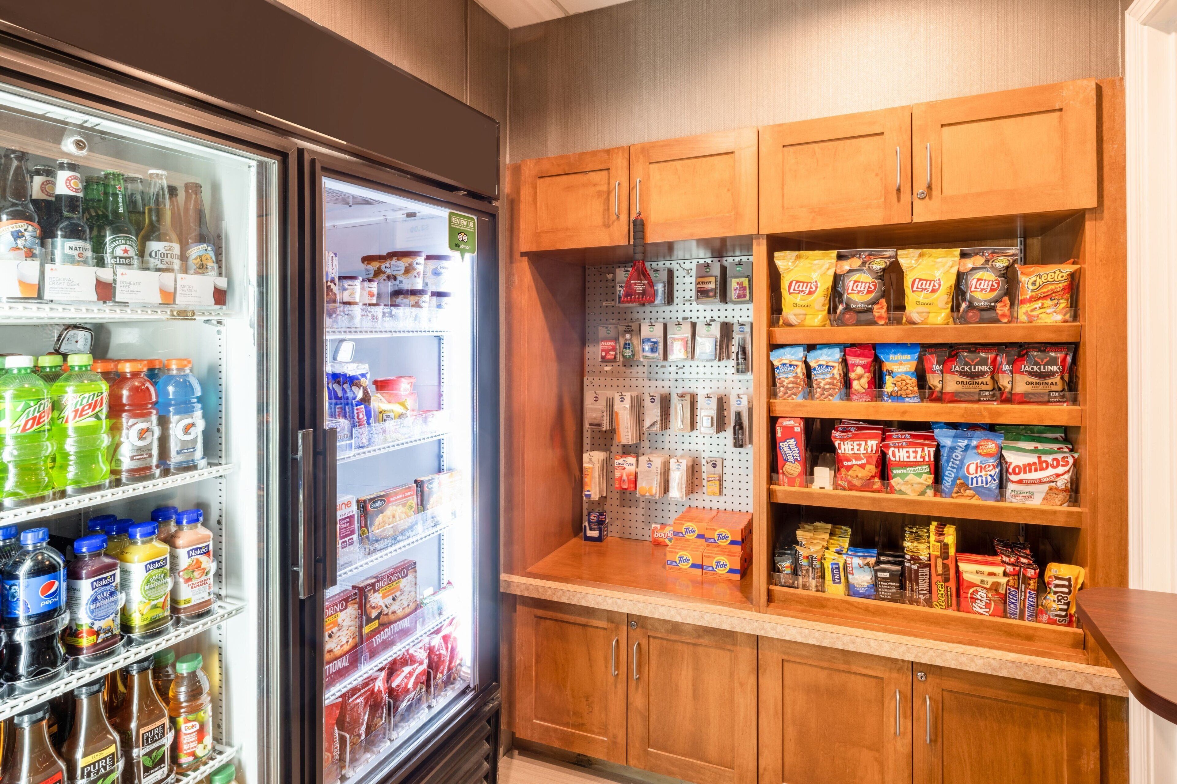 A kitchenette with a refrigerator stocked with beverages, a microwave, and wooden cabinets containing snacks and condiments.