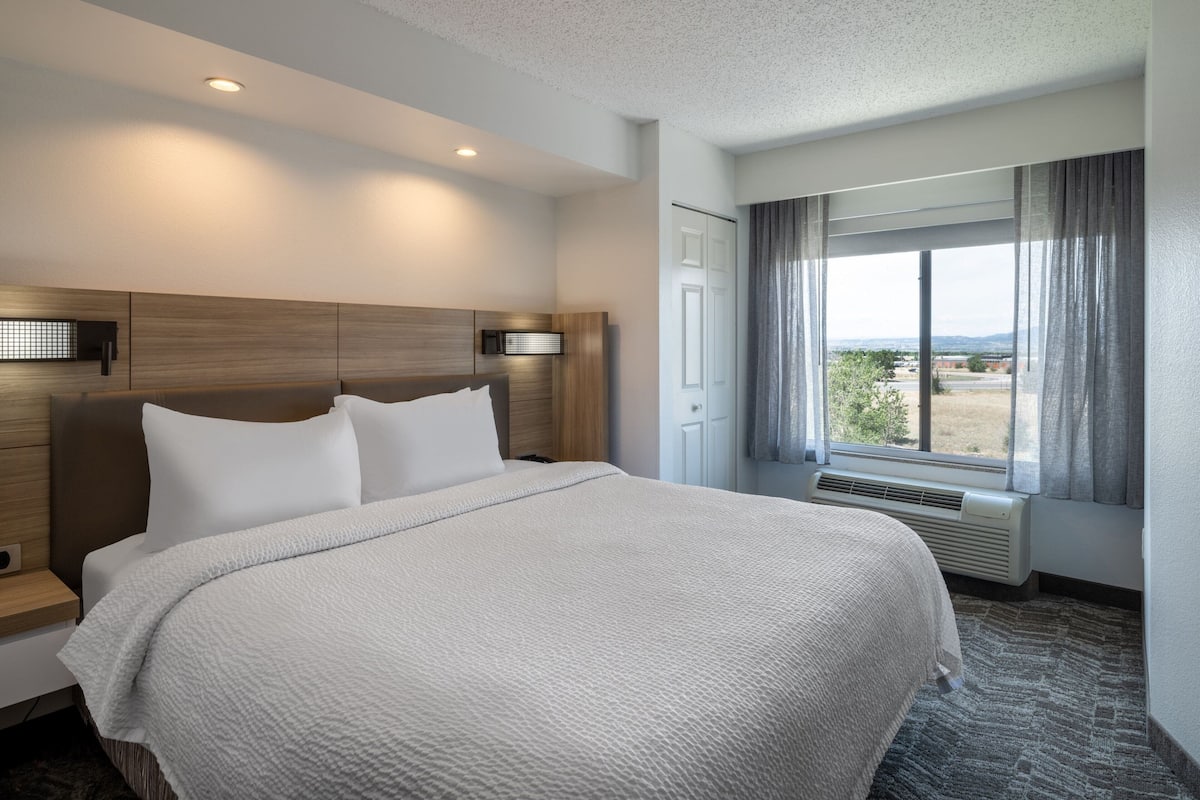 A hotel room with a large bed, a wooden headboard, a window with curtains, and a view of the outdoors.