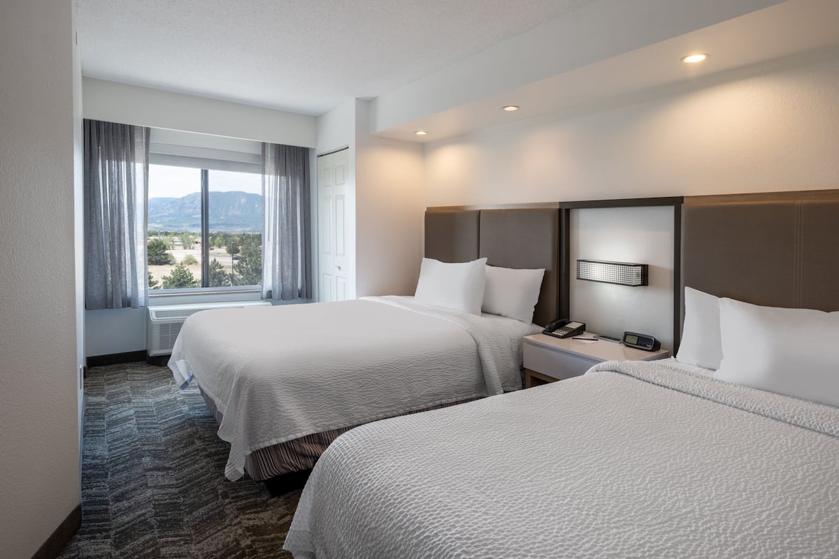 A hotel room with two beds, a large window with curtains, a nightstand with a phone, and a view of the outdoors.