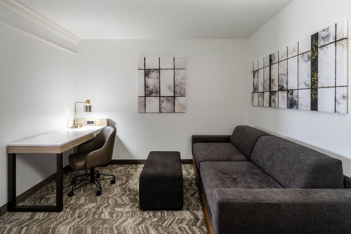 A modern living room with a desk, a grey sofa, a black ottoman, and abstract wall art.