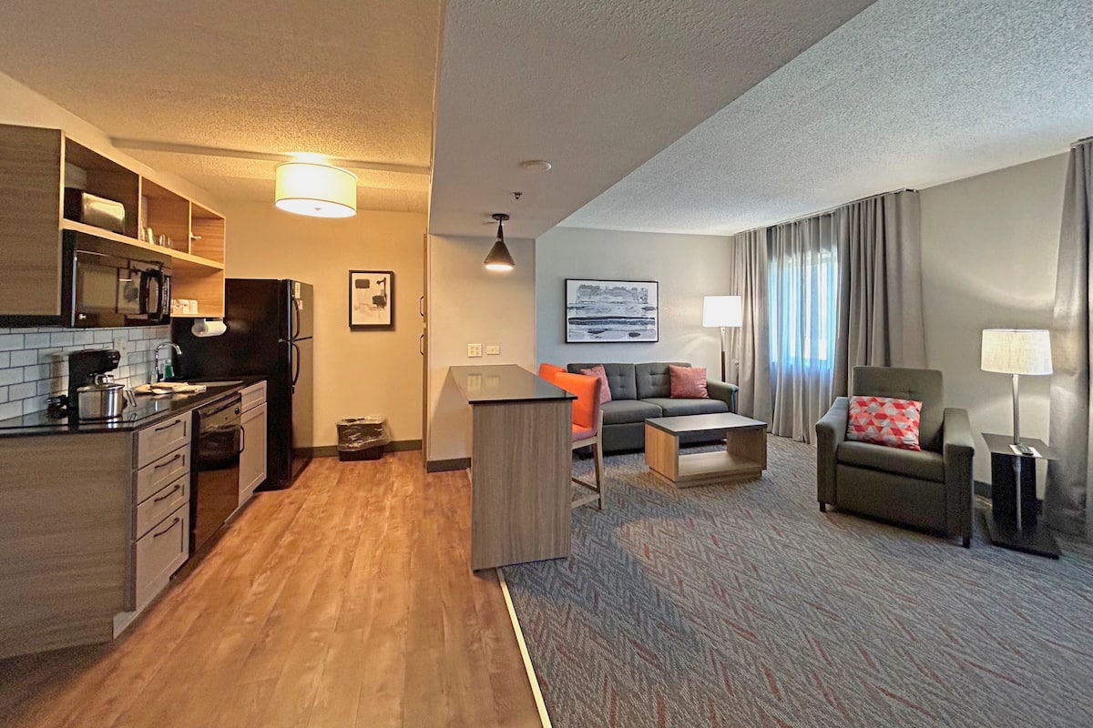 Una habitación de hotel compacta con kitchenette, un sofá, una mesita y una silla.