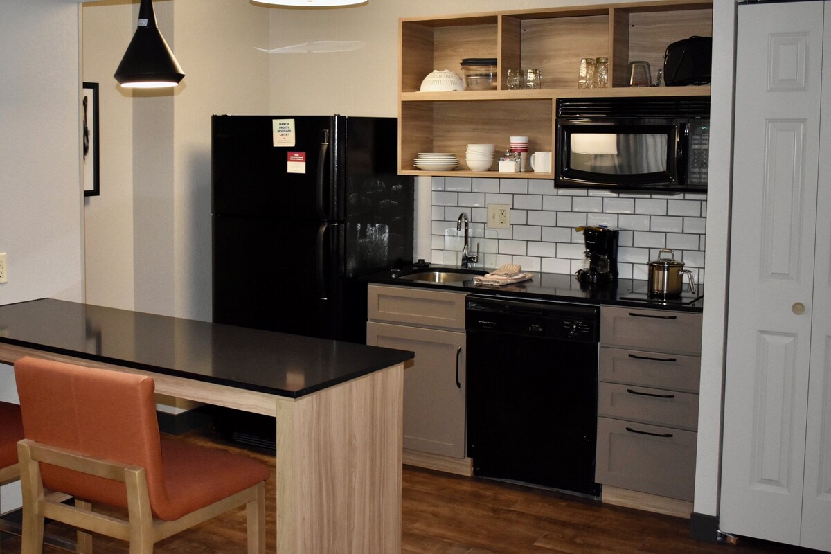 Una cocina compacta con un refrigerador negro, un microondas, una cafetera y un lavavajillas. Hay un pequeño comedor con dos sillas y una mesa.