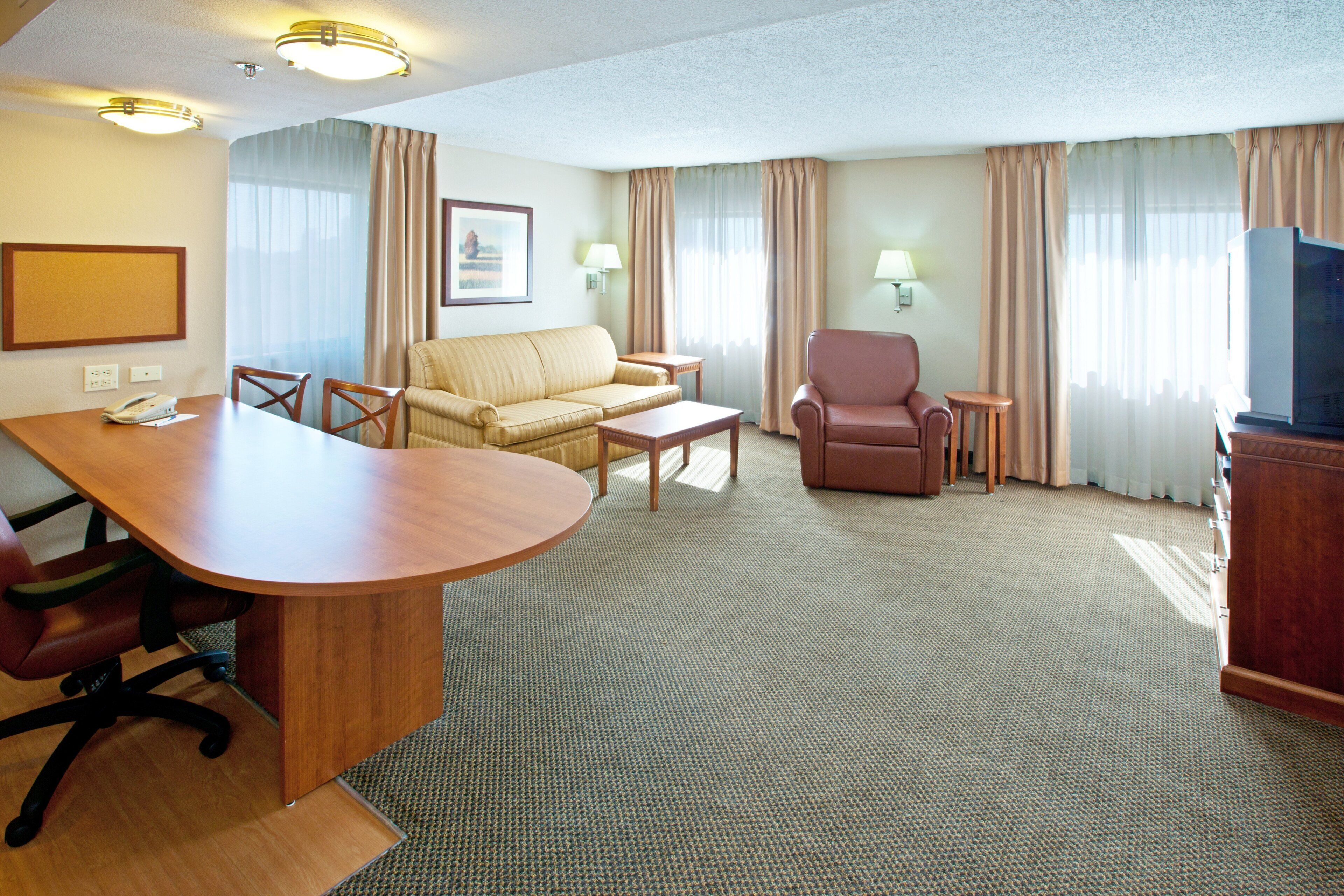 A hotel room with a wooden desk, two armchairs, a sofa, a television, and a framed picture on the wall.