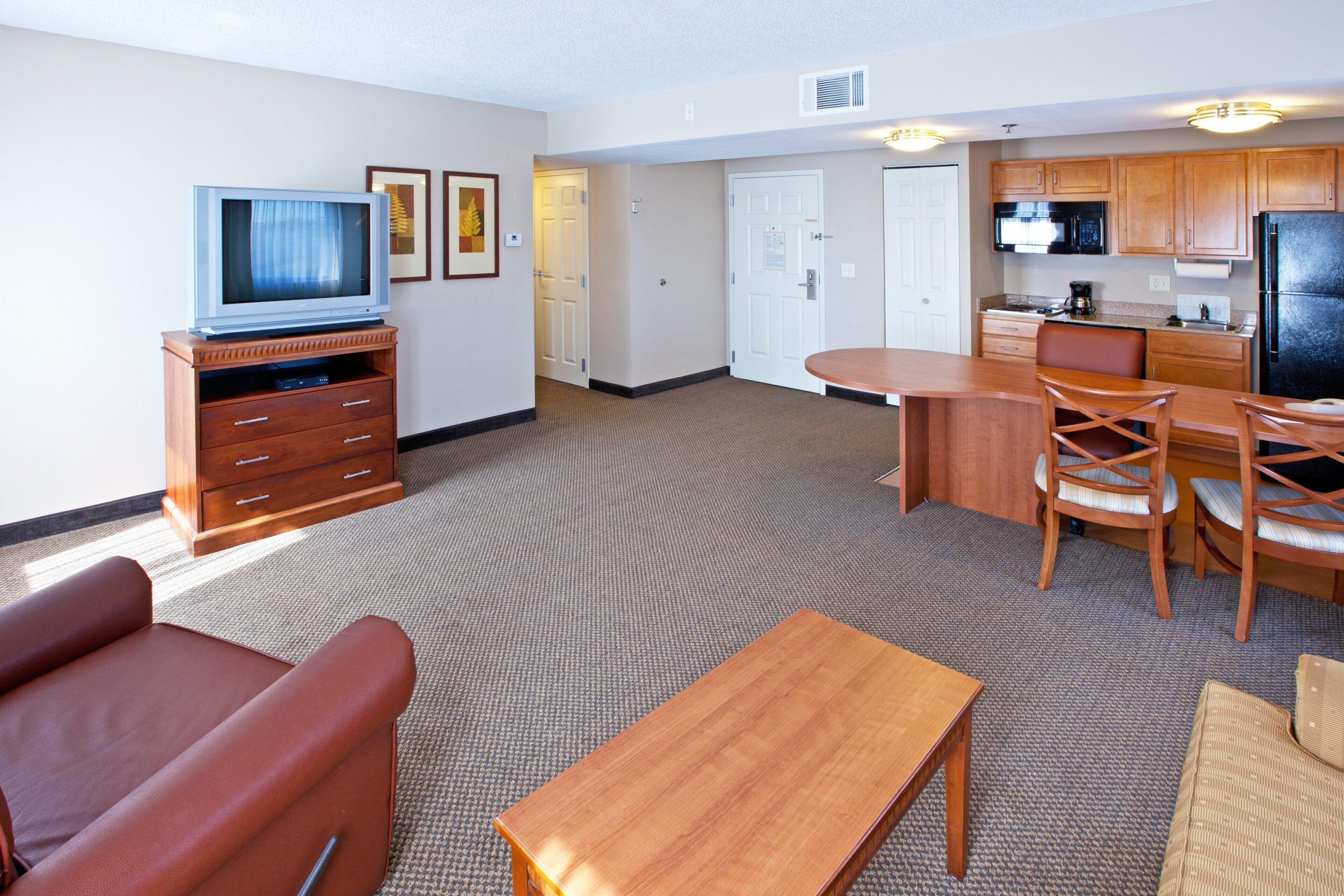A hotel room with a television, a dining table with chairs, a sofa, and a kitchen area with a refrigerator and microwave.
