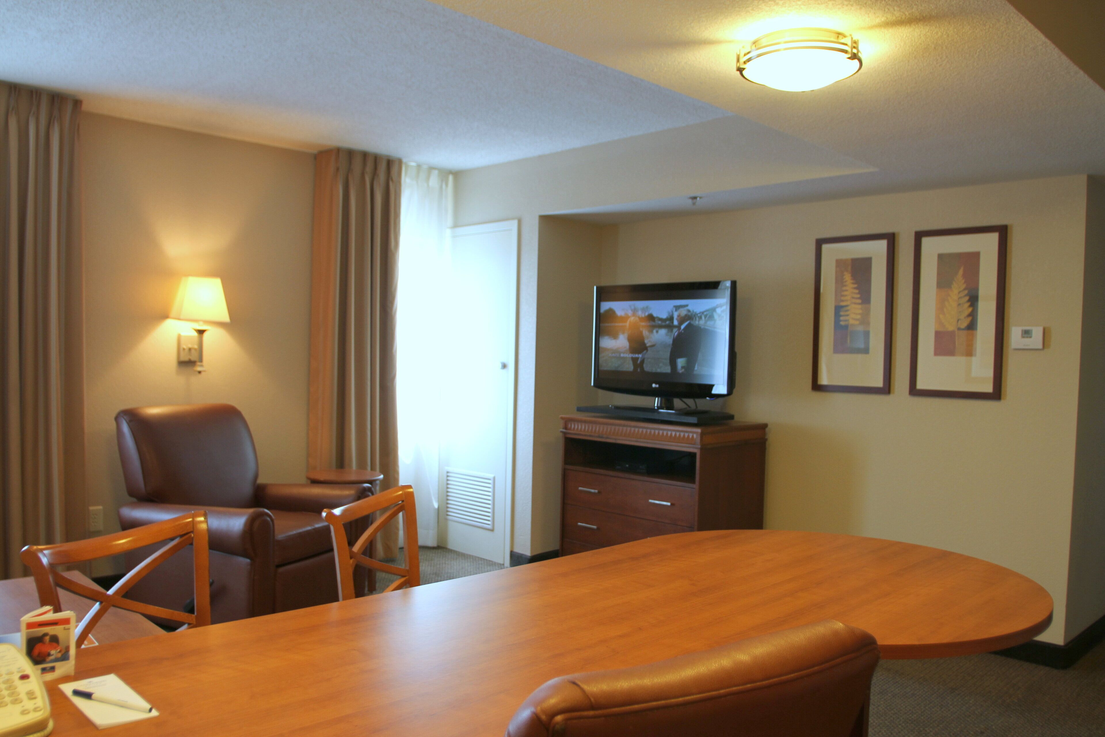 Ein Hotelzimmer mit einem Holzespeisetisch, zwei Ledersesseln, einem Fernseher auf einem Holzständer und einer Wandleuchte.