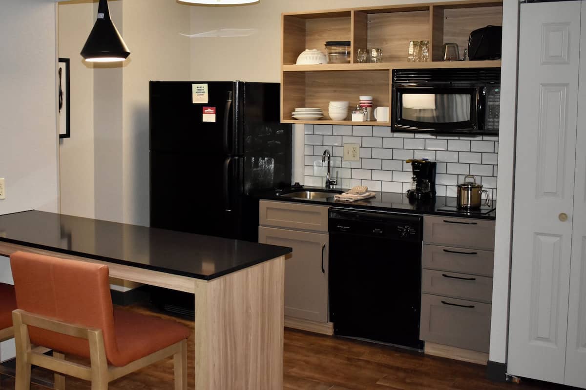 Una cocina compacta con un refrigerador negro, un microondas, una cafetera y un lavavajillas. Hay un pequeño comedor con dos sillas y una mesa.