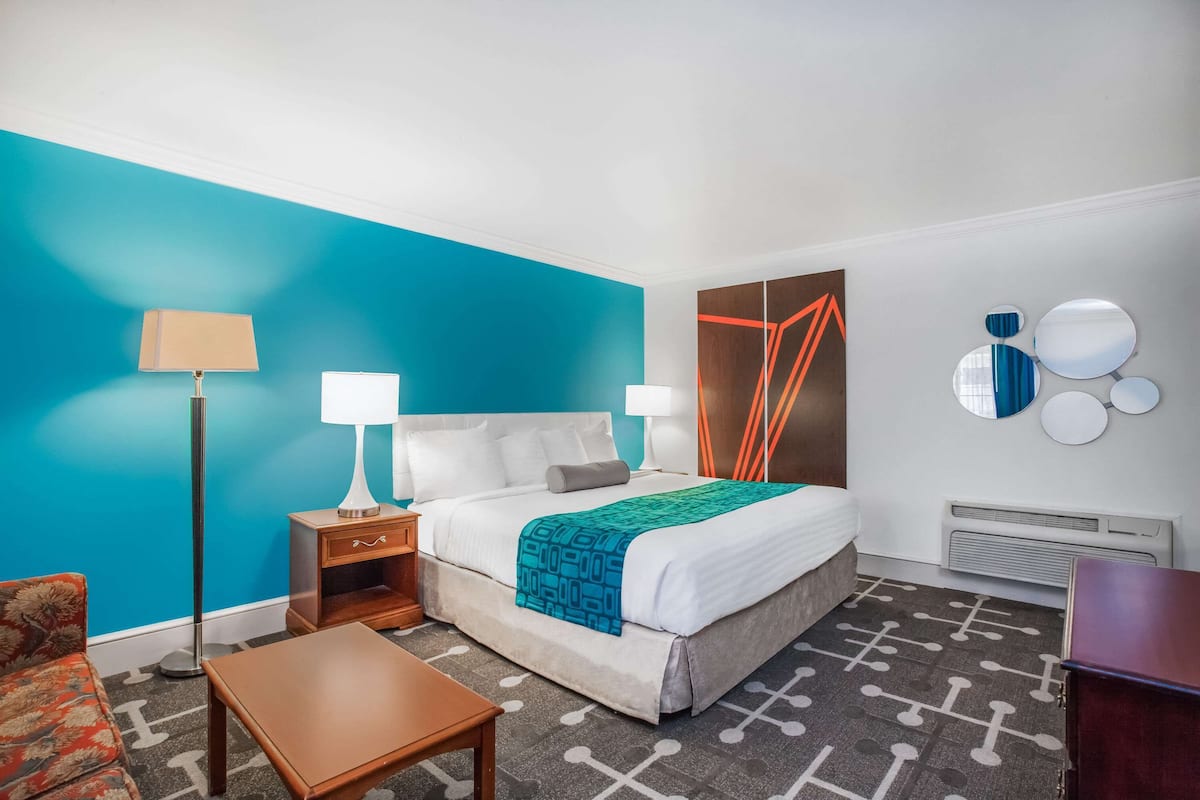 Ein Hotelzimmer mit einem großen Bett, zwei Nachttischen mit Lampen, einem kleinen Holztisch, einem gemusterten Teppich und einer Wand mit Dekorationselementen aus Spiegeln.