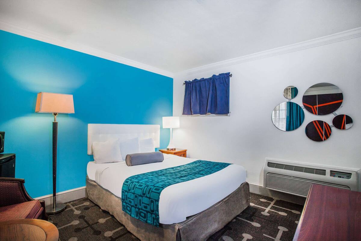 Ein Hotelzimmer mit einem großen Bett, einem Schreibtisch, einem Stuhl, einer Lampe, einem Fenster mit Vorhängen und Wanddekoration.