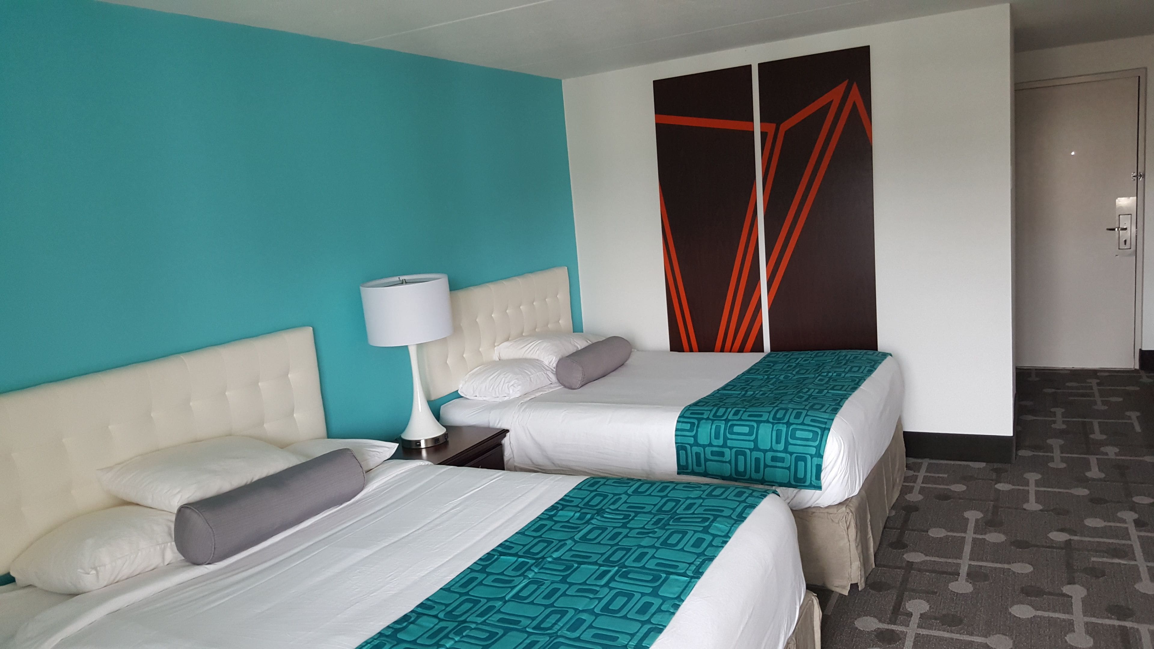 A hotel room with two beds, a white headboard, a teal accent wall, a patterned bedspread, and a nightstand with a lamp.