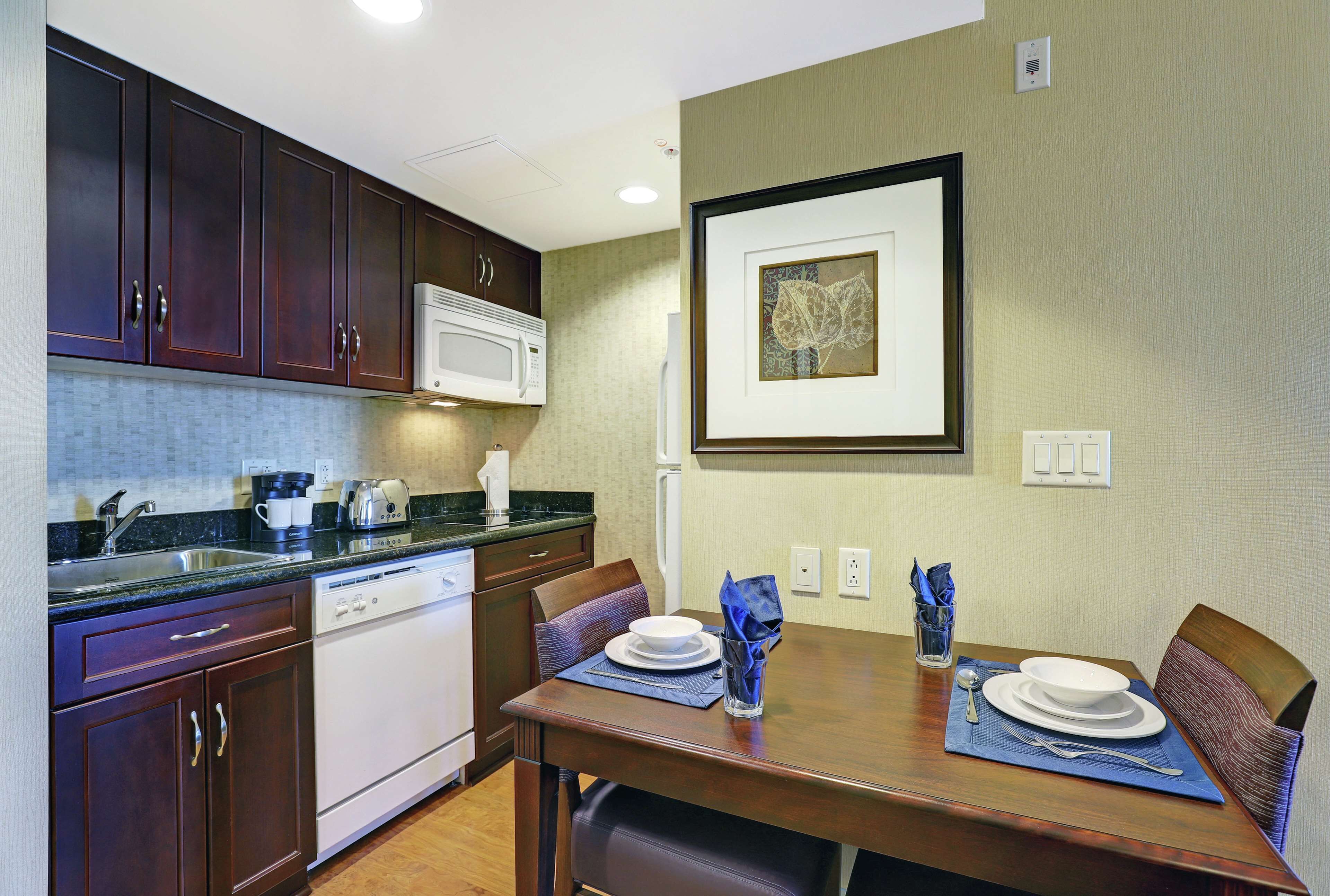 A compact kitchen with dark wooden cabinets, a white microwave, and a dining area with a wooden table set for two.
