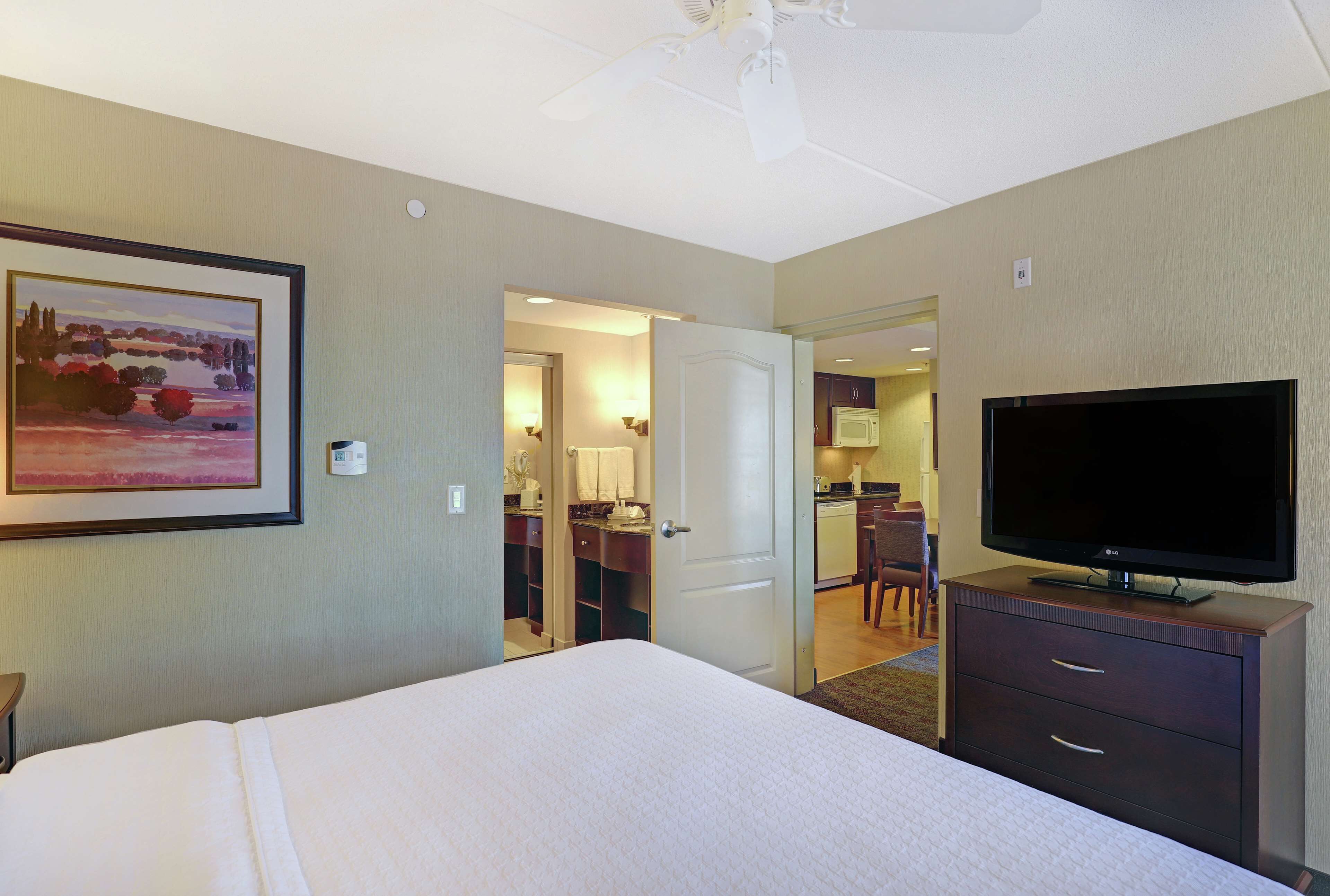 A hotel room with a bed, a television, a painting, and a kitchenette in the background.