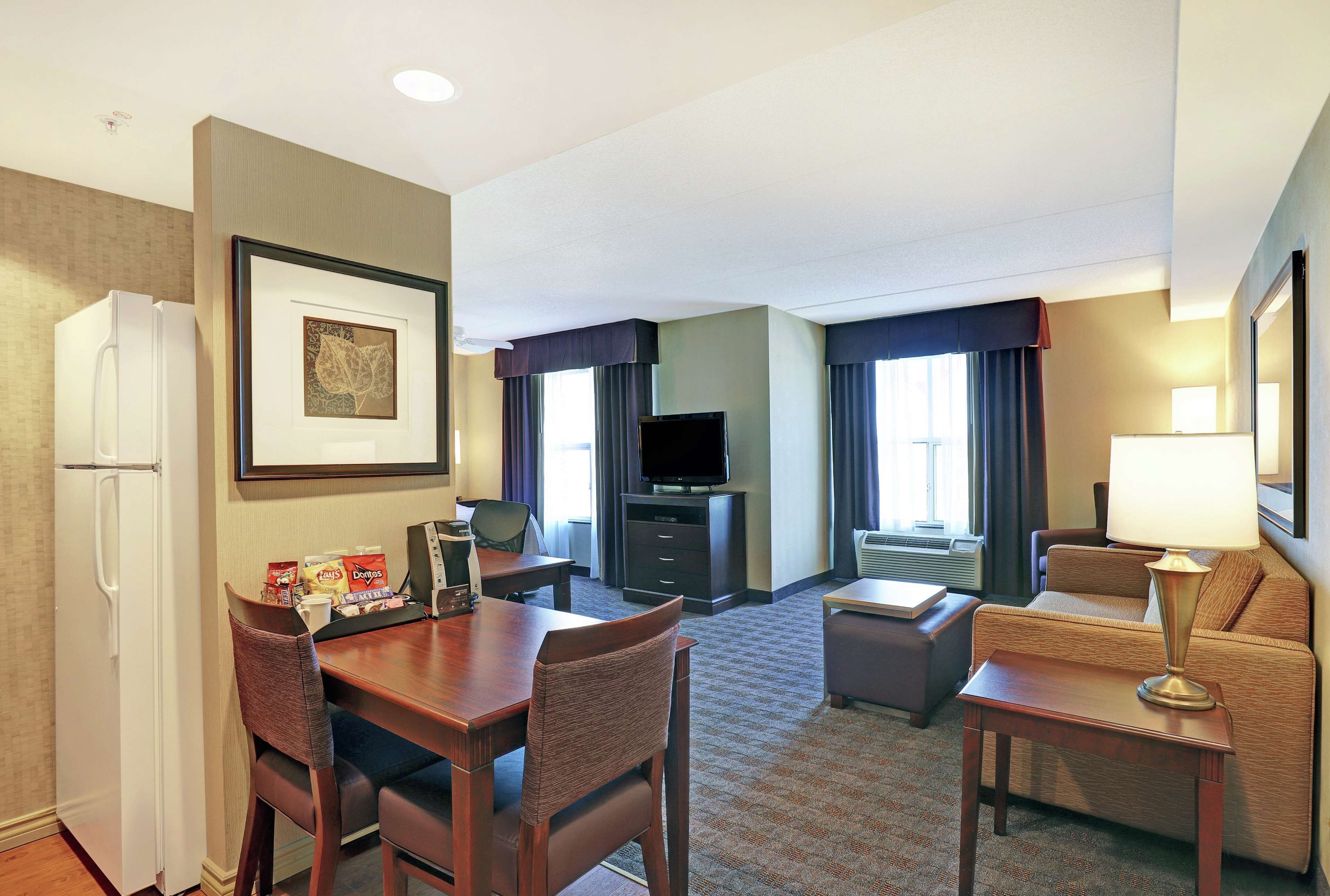 Habitación de hotel con mesa de comedor, sillas, refrigerador, televisor, sofá y lámpara.