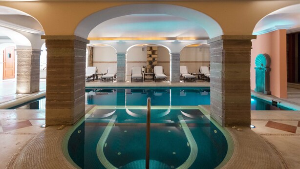 Una piscina interior con un poste central, rodeada de sillones de descanso y pilares de piedra.