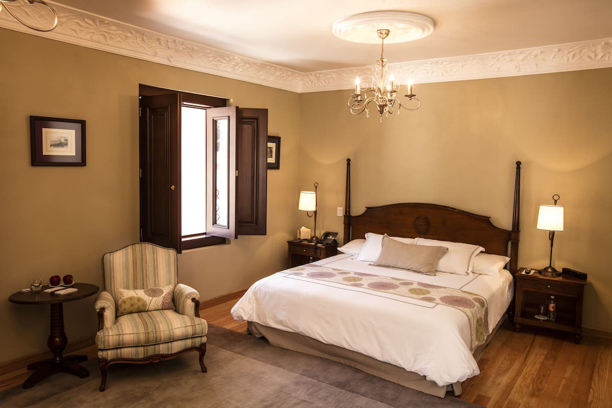 Un dormitorio con una cama grande, una silla, una mesita de noche, una lámpara y una ventana con contraventanas abiertas.