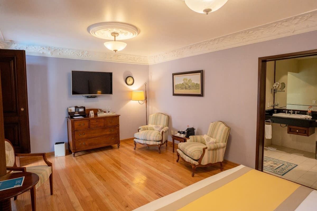 Una habitación con piso de madera, televisor, reloj, dos sillones, cómoda, lámpara y baño con lavamanos y espejo.