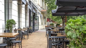 Un café al aire libre con mesas y sillas, una terraza de madera y un edificio con ventanas amplias y un balcón.
