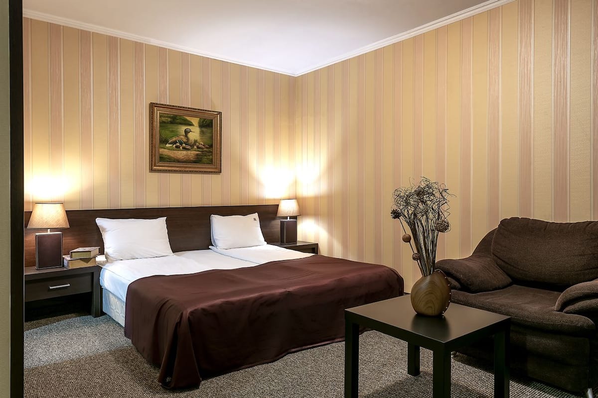 A hotel room with a bed, two bedside tables with lamps, a brown sofa, a small table with a vase, and a painting on the wall.