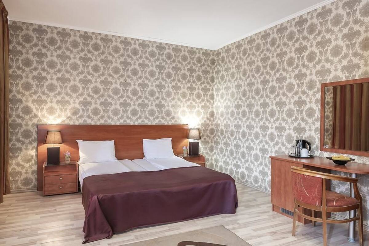 A hotel room with a large bed, a wooden nightstand with a lamp, a desk with a mirror, and patterned wallpaper.