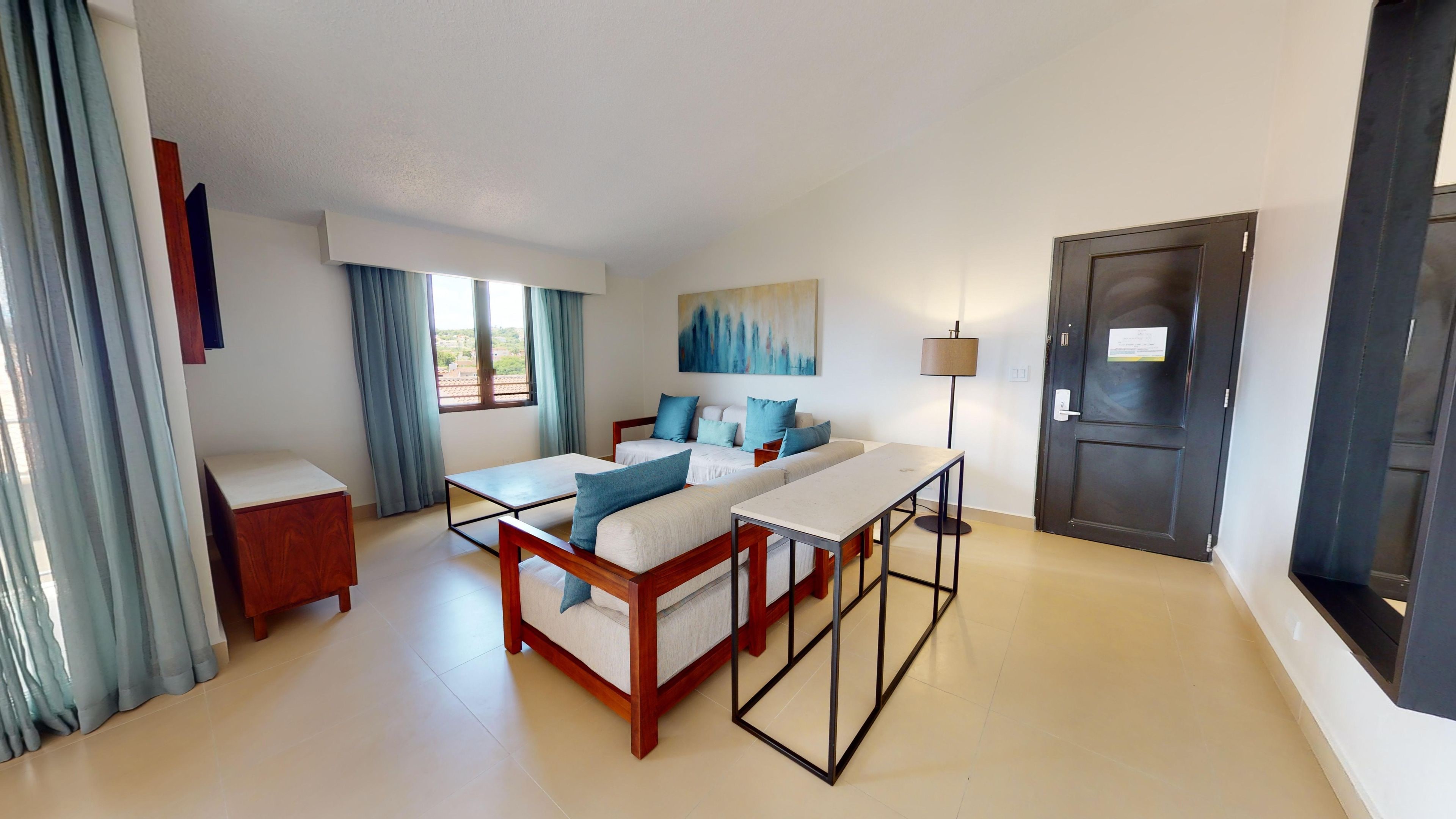 A hotel room with a bed, a desk, a chair, a lamp, and a door.