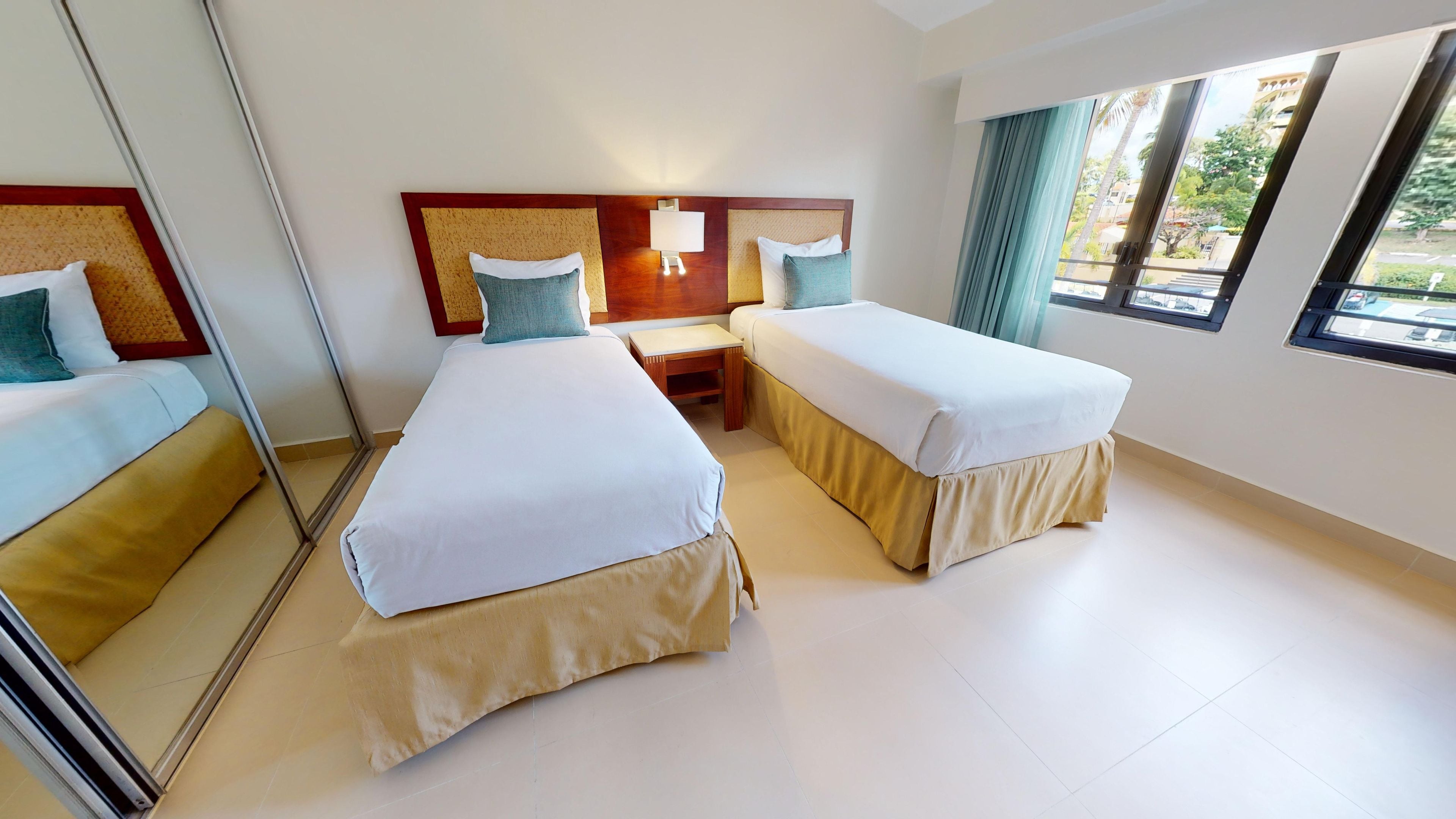 Une chambre d’hôtel avec deux lits, une petite table, un miroir et une fenêtre donnant sur des palmiers.