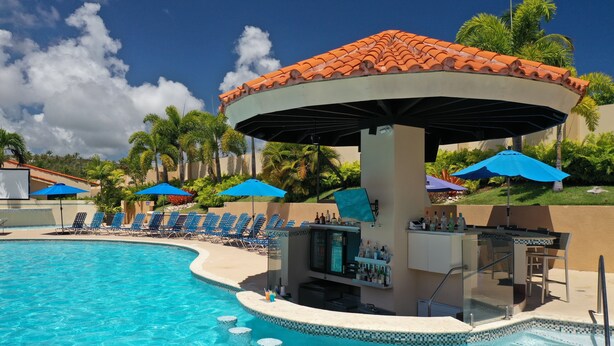 Un bar situé au bord de la piscine, doté d’un toit en tuiles, de parasols bleus et de chaises longues.