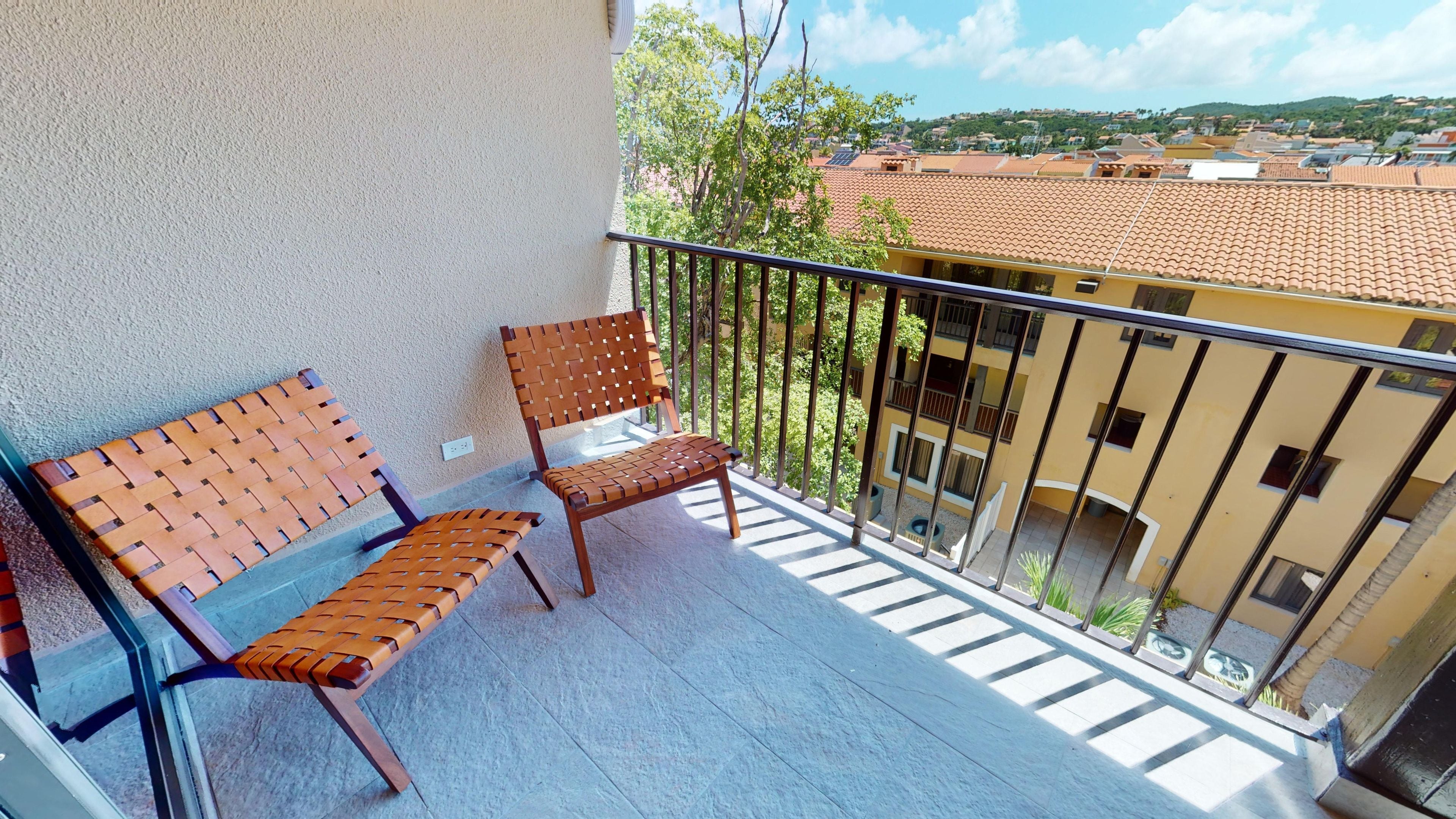 Varanda com duas cadeiras de madeira e um banco, com vista para uma área residencial com telhados de terracota.