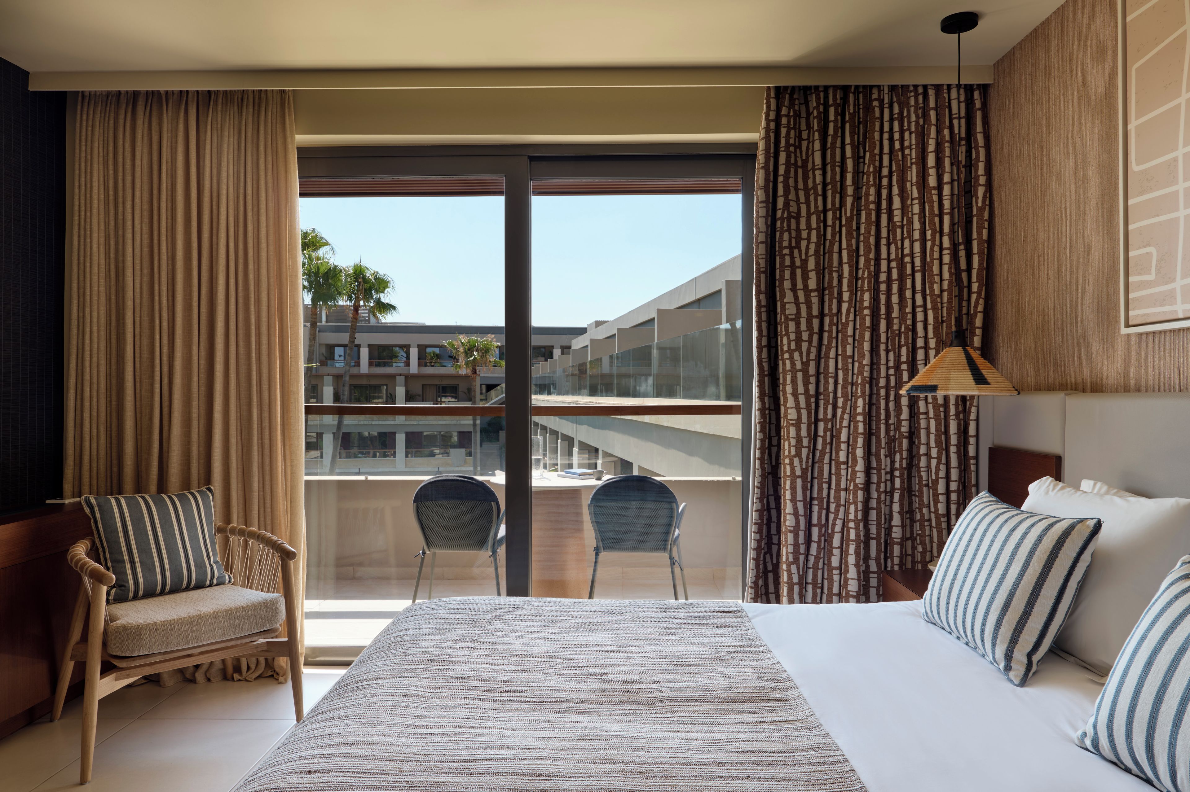 A hotel room with a large bed, two chairs, a view of a building through a glass door, and a patterned curtain.