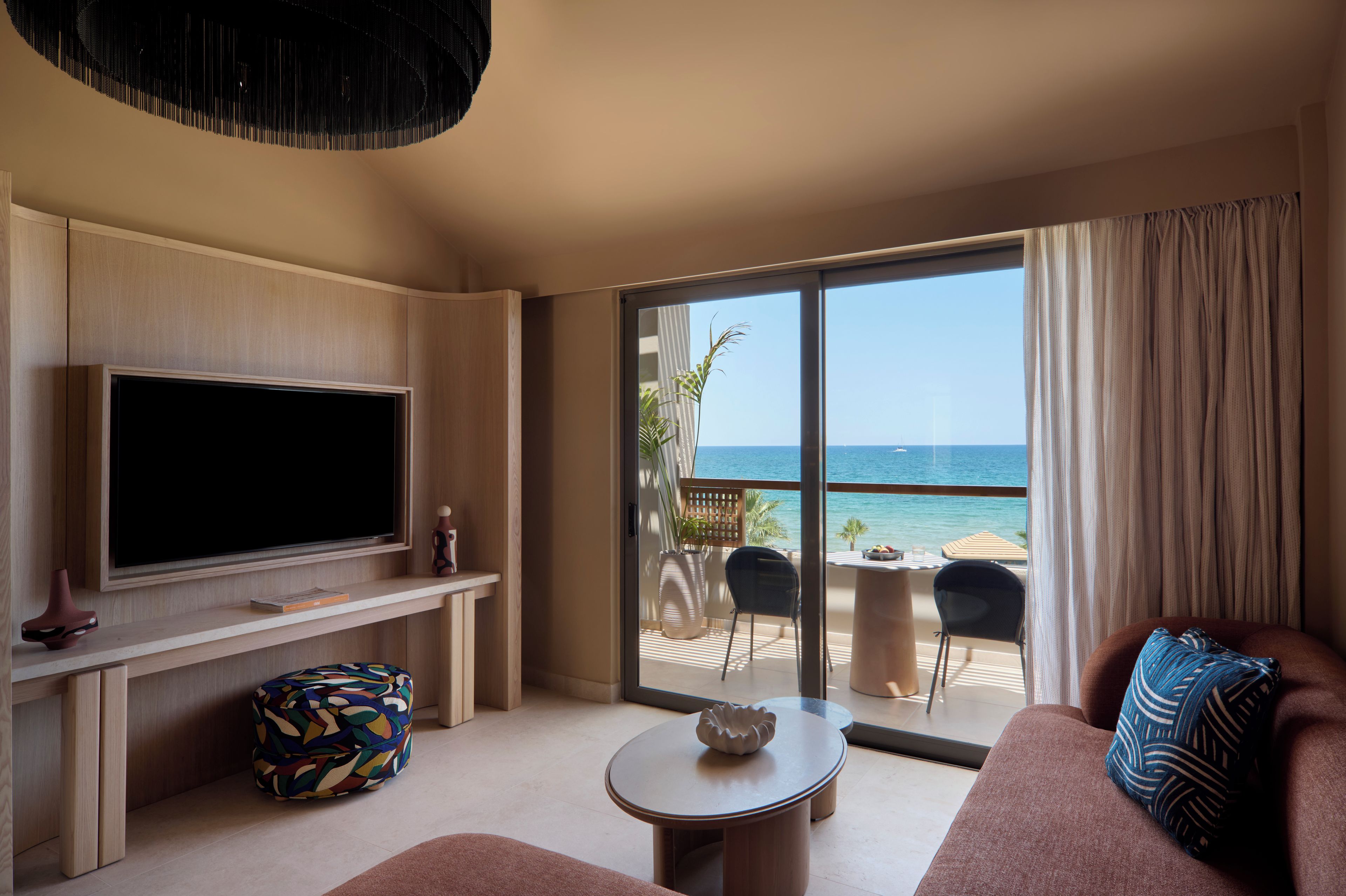 A modern living room with a flat-screen TV, a coffee table, and a sofa. There is a balcony with a view of the sea and a dining area with chairs and a table.