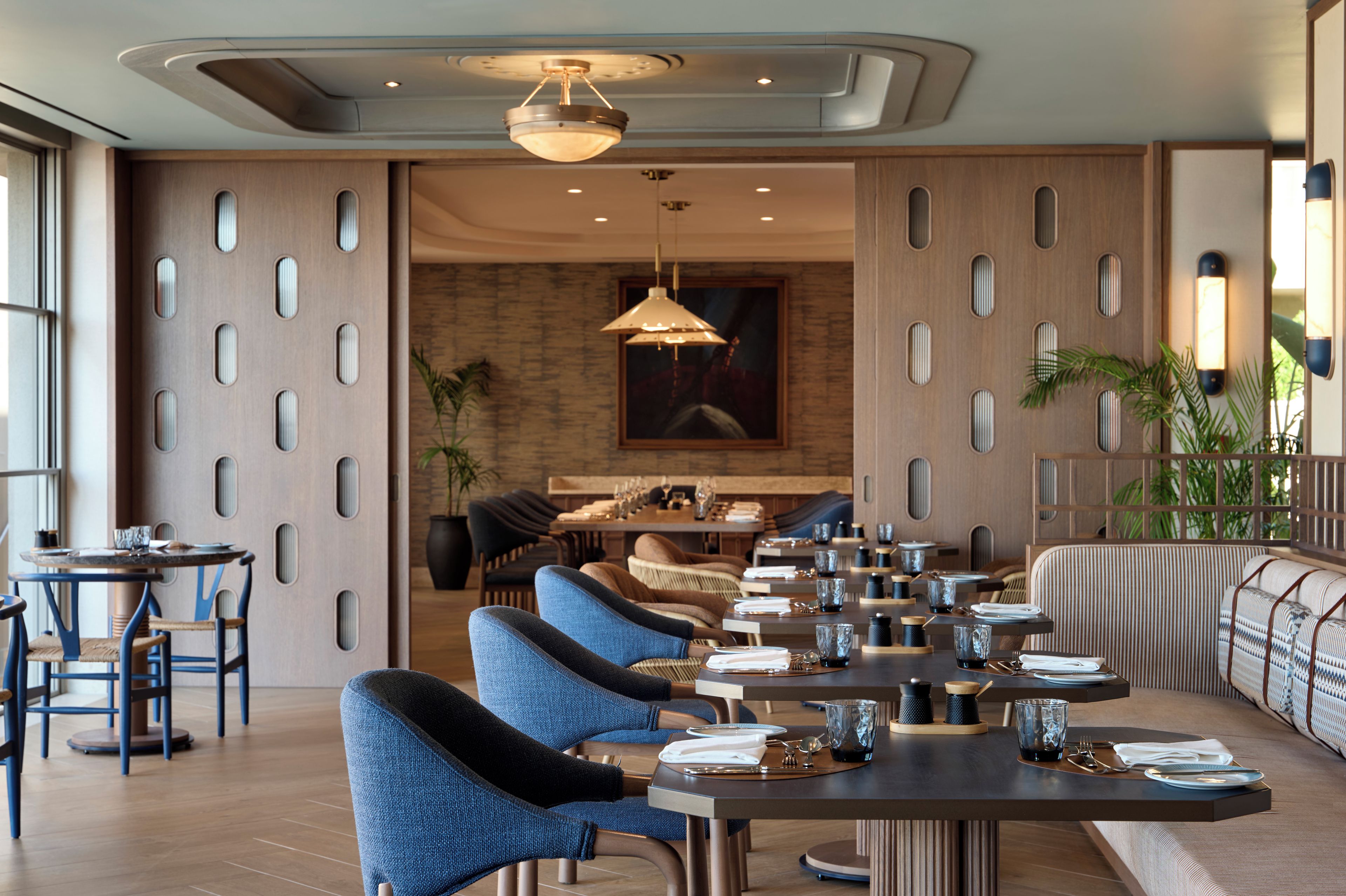 Een moderne eetruimte met een lange tafel, blauwe fauteuils en een eigentijds plafondontwerp.