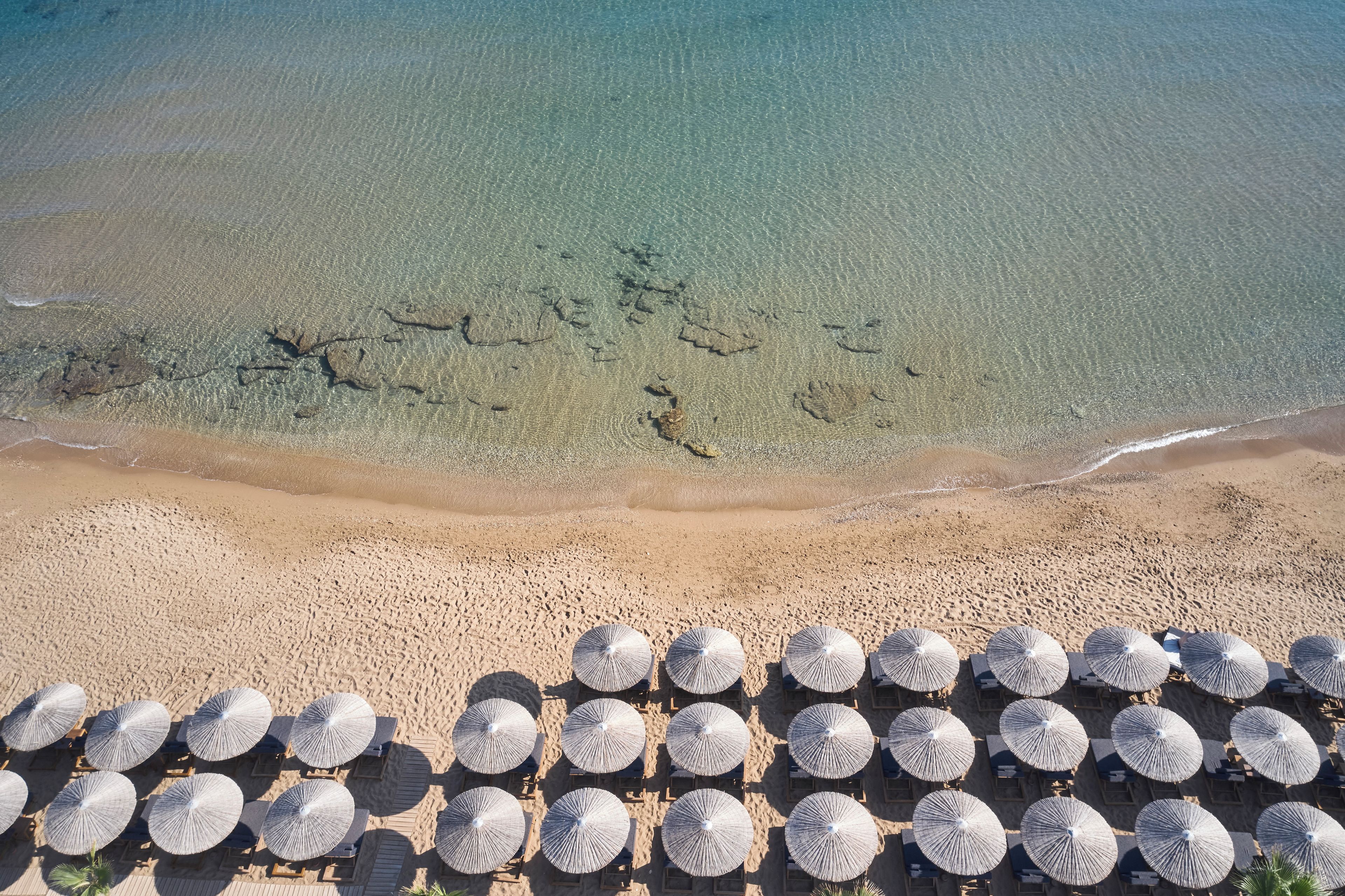 An aerial view of a beach with clear water, a sandy shore, and rows of sun umbrellas and lounge chairs.