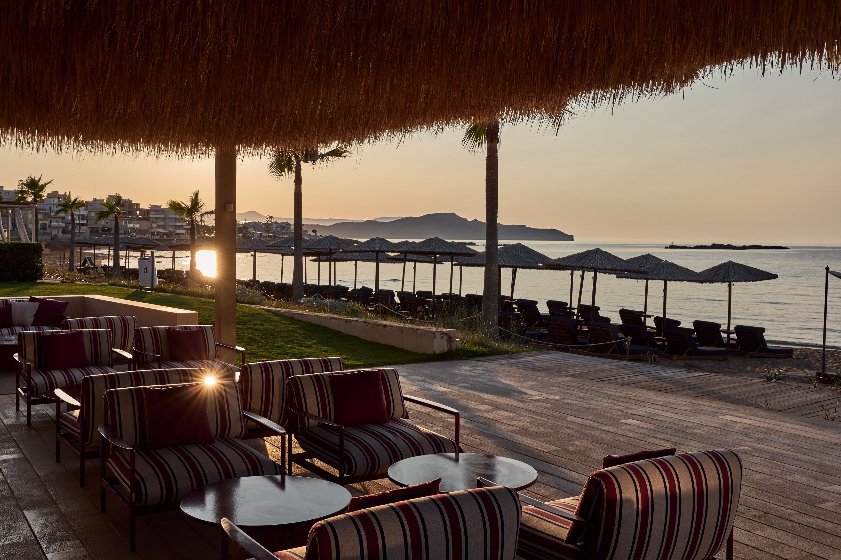 A seaside patio with striped furniture, umbrellas, and a view of the ocean at sunset.