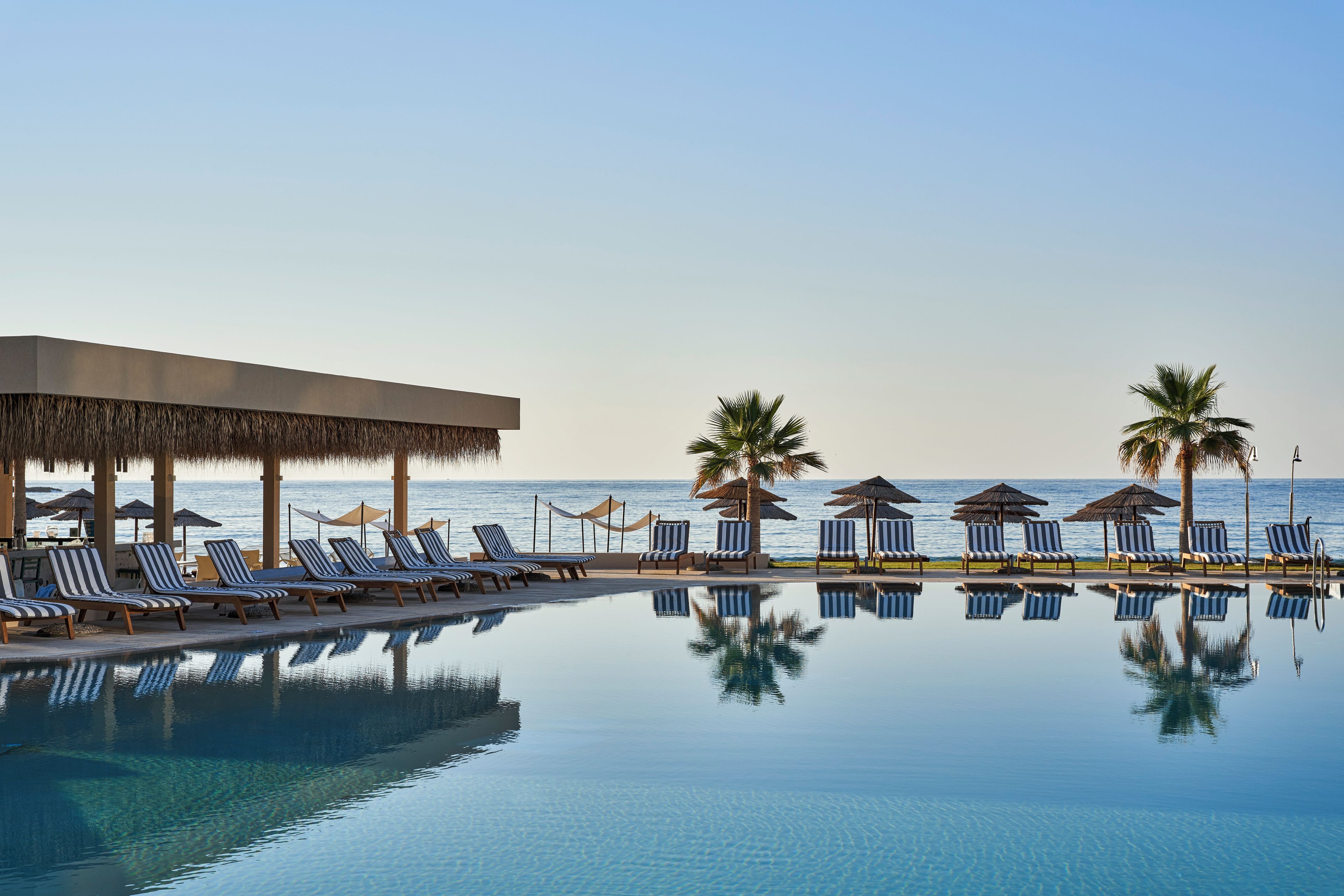 A poolside area with lounge chairs and umbrellas, a clear view of the ocean, and a reflection in the pool.
