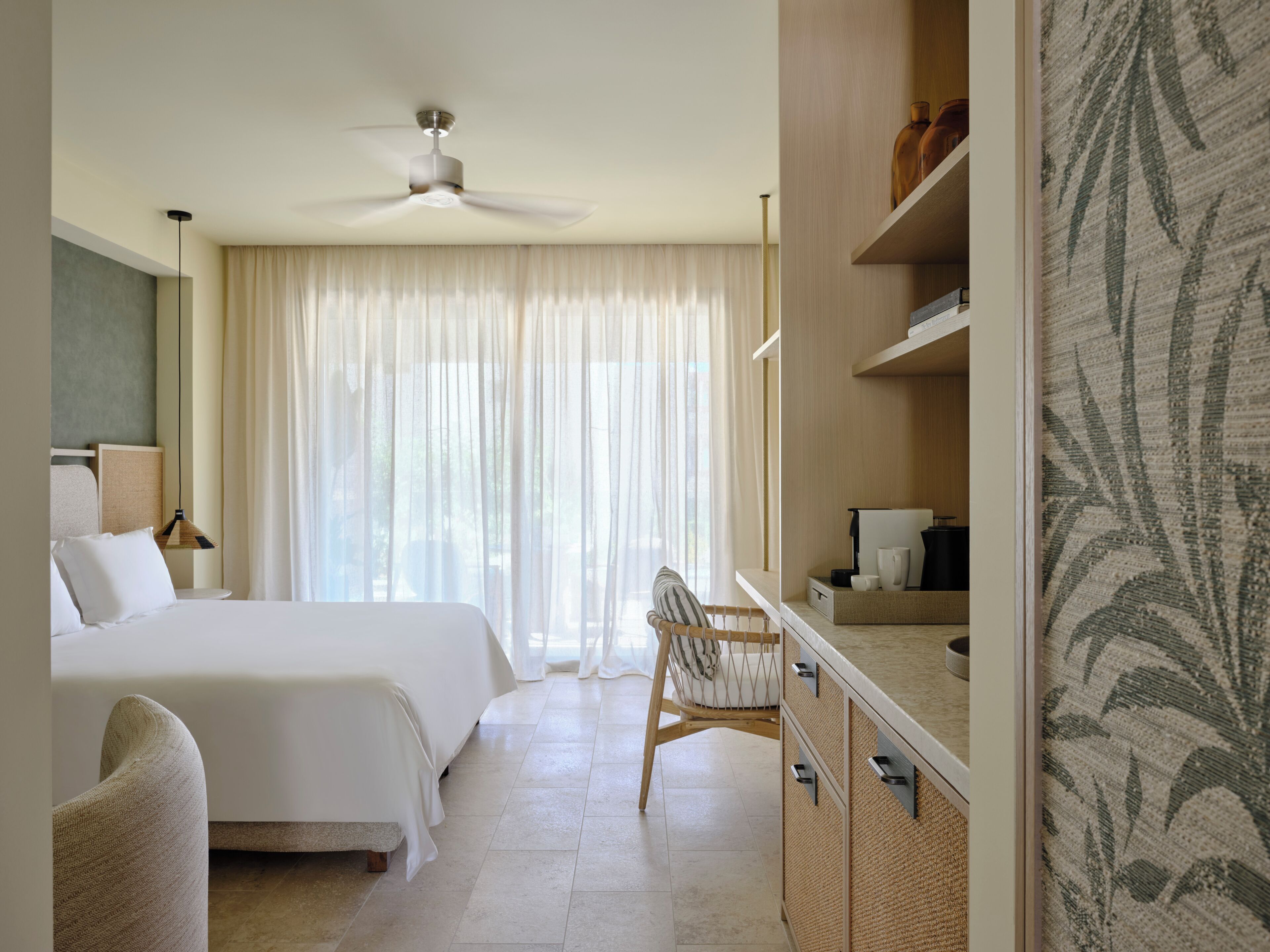 A hotel room with a bed, a chair, a desk with a mirror, and a ceiling fan.