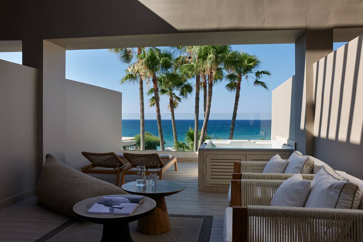 Un área lounge moderna al aire libre con mobiliario de mimbre, una mesa redonda de madera y vista al océano y palmeras.