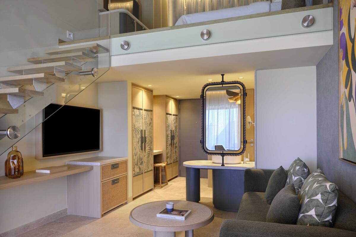 Una sala de estar moderna con un televisor de pantalla plana, una mesa redonda de centro, un sofá con cojines estampados, una escalera y un espejo grande.