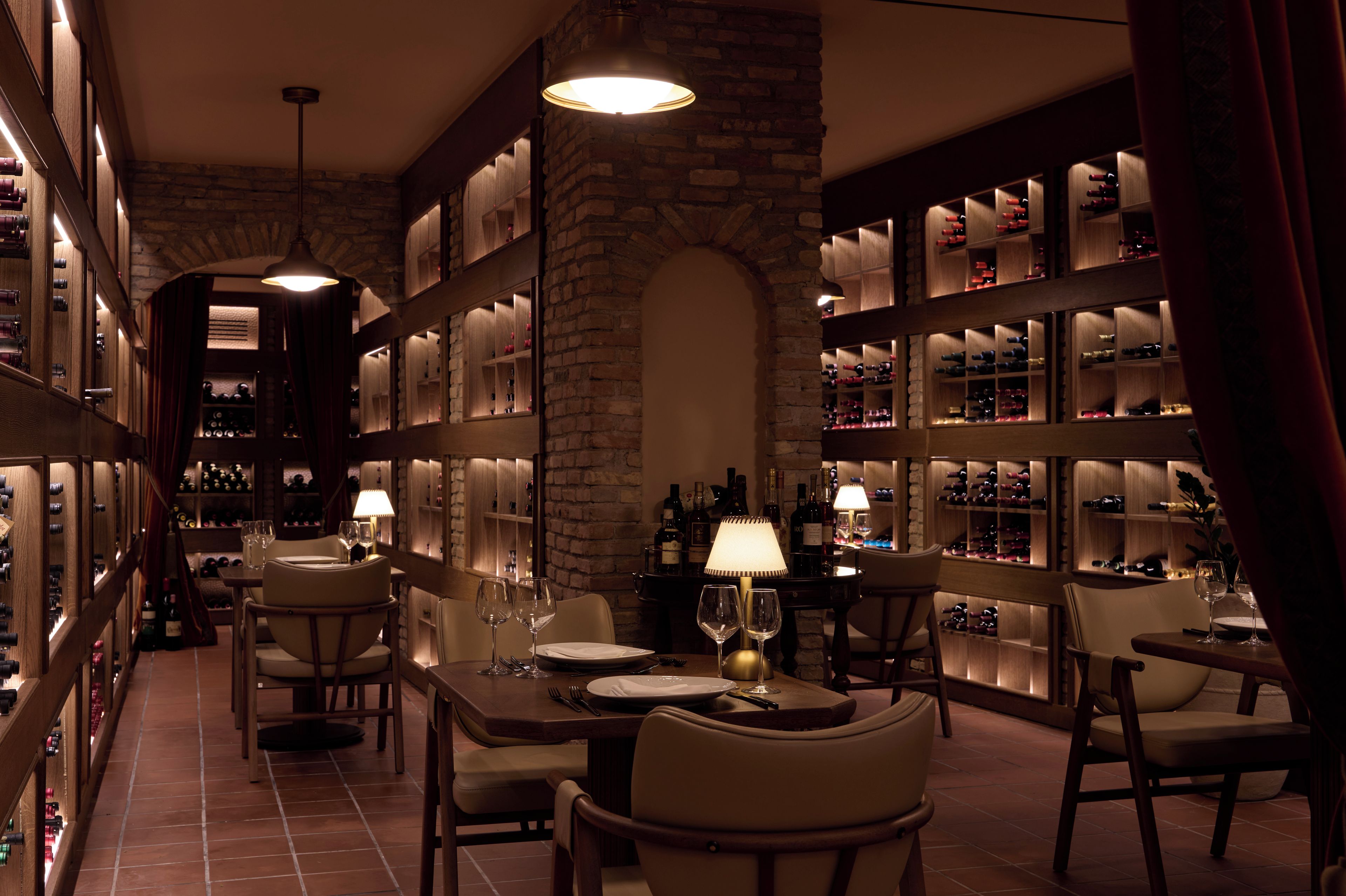 A restaurant with a brick wall, wooden shelves with wine bottles, and tables set for dining.