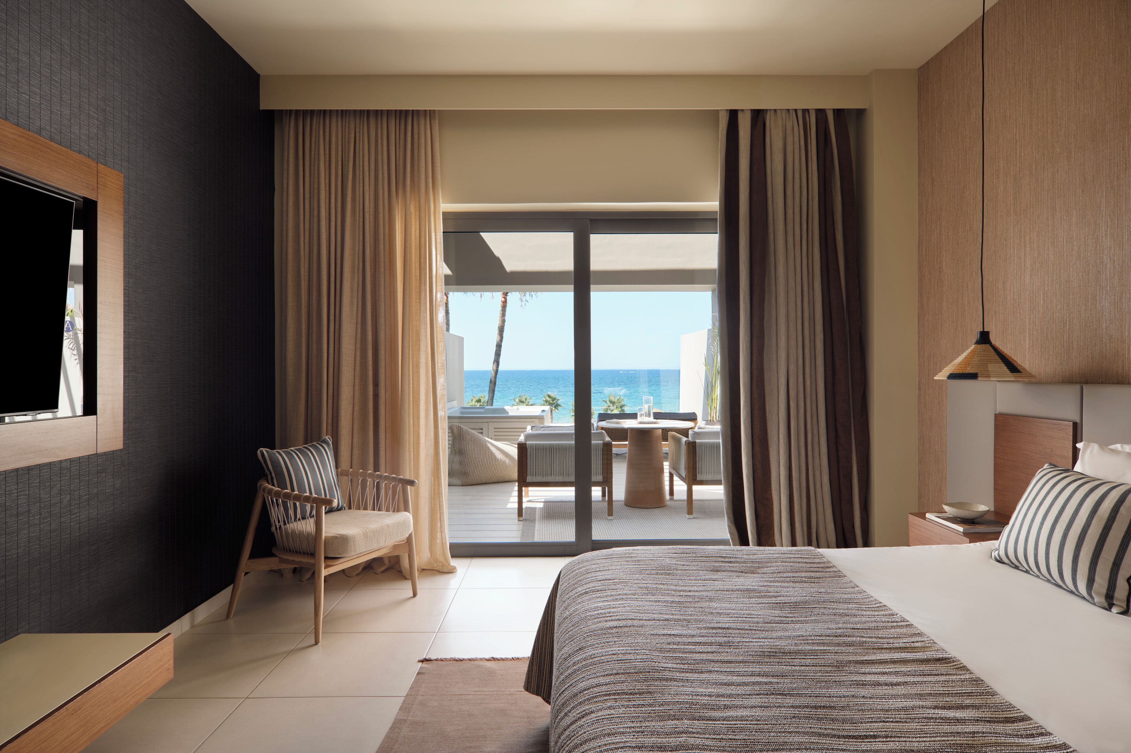 A hotel room with a bed, a chair, a table, and a view of the sea.