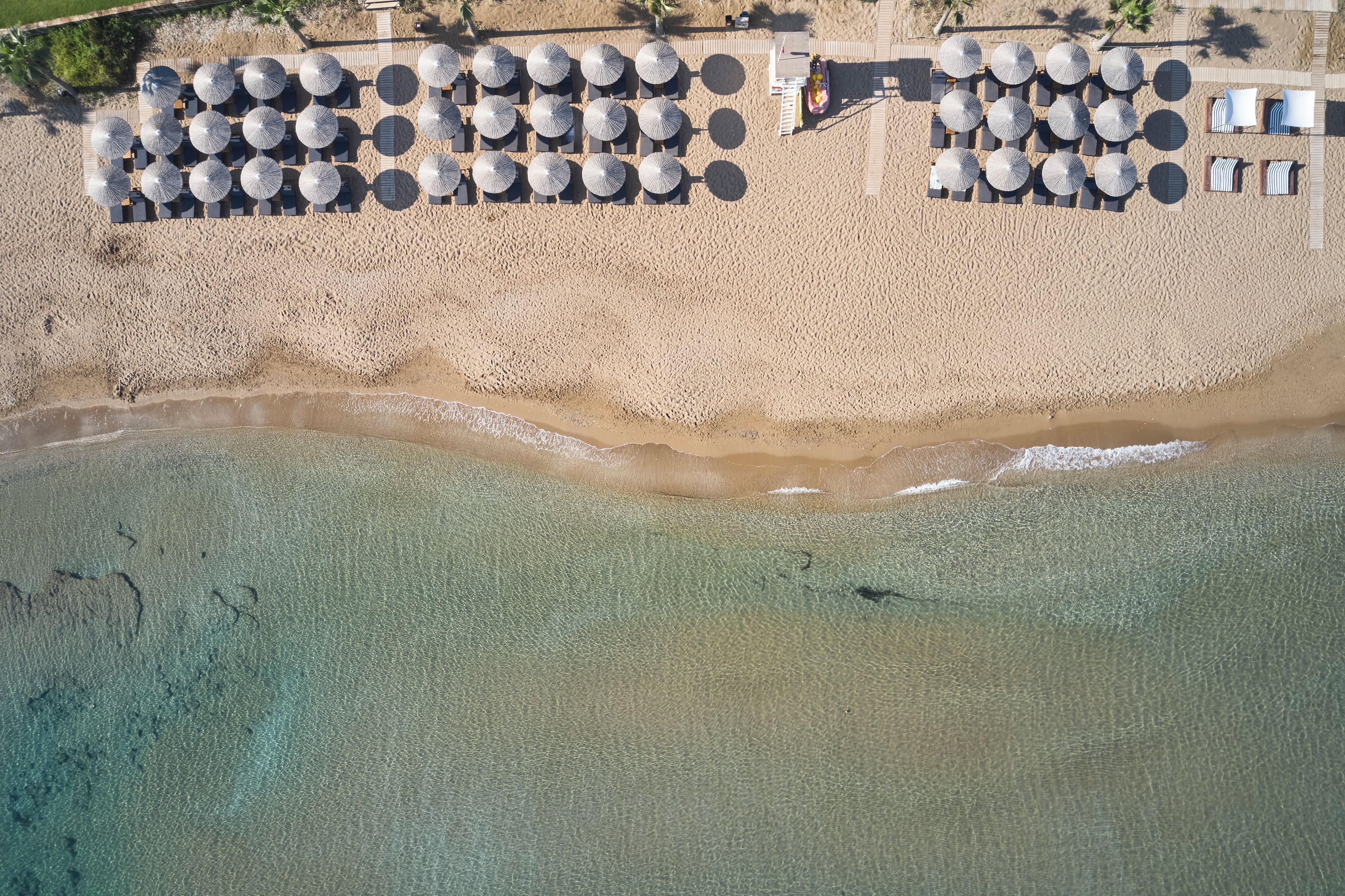 An aerial view of a beach with rows of sun umbrellas and lounge chairs, a wooden walkway, and clear turquoise water.